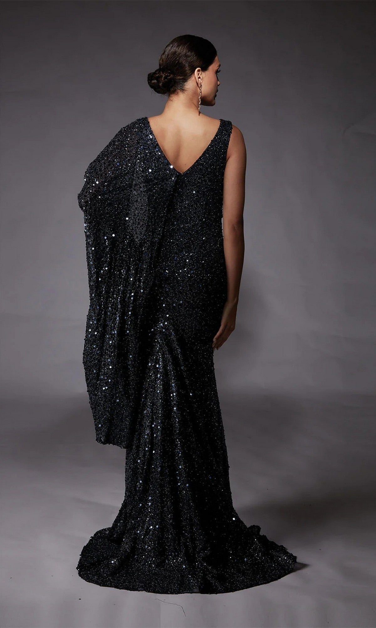 A woman faces away, wearing the shimmering Alyce 27772 long formal dress—a black sequin-beaded gown with a draped design and V-shaped back—standing against a plain gray background.